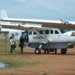 Small aircraft landing on remote airstrip with safari vehicles and wildlife in the background